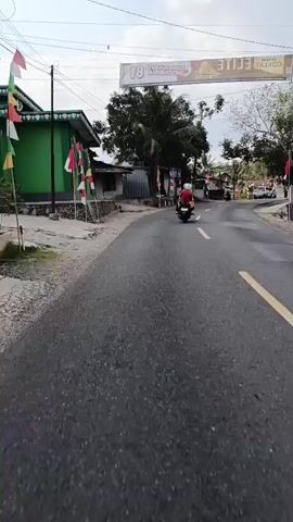 The video shows a motorcycle with a person in a red shirt riding on a paved road. There are trees and buildings on the sides of the road. Several Indonesian flags are displayed along the road. The sky is cloudy.