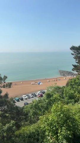 The video captures an elevated view of a beach, showcasing a serene coastal scene on a clear, sunny day. The beach, composed of light brown sand, is sparsely populated with people sunbathing, walking, or setting up beach tents. The calm ocean stretches out to the horizon under a cloudless, light blue sky. A rock structure extends into the water on the right side of the beach, acting as a breakwater. Adjacent to the beach is a parking lot filled with cars. Lush green trees and shrubs frame the lower portion of the view, partially obscuring the parking lot and providing a natural foreground to the scene.