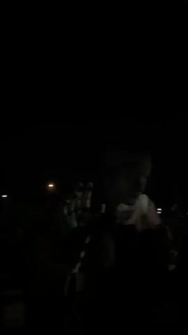 The video captures a night-time outdoor concert or festival. It begins with a brief, close-up shot of a performer, whose face and hand are dimly illuminated, appearing to be wearing light-colored attire or makeup. The camera then pans up to reveal the stage and audience from a distance. The scene is very dark, with the only significant illumination coming from the stage lights and laser beams. Bright white stage lights are visible across the top of the stage structure. Throughout the latter part of the video, vibrant green laser lights sweep across the dark sky, adding to the concert atmosphere. The audience is largely obscured in silhouette against the bright stage lights. The event appears to be taking place in the Sdot Dan Regional Council, Israel.
