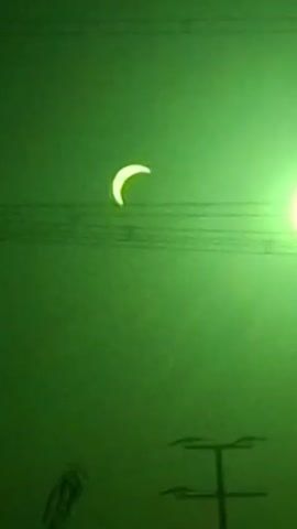The video shows a solar eclipse with the moon partially obscuring the sun. The sky is a deep green hue, possibly due to the atmospheric conditions during the eclipse. There are visible power lines and a utility pole in the foreground, suggesting an outdoor setting. The sun, filtered by the eclipse, appears as a bright crescent shape against the dark sky. The video is of poor quality, with motion blur and graininess, making it difficult to discern finer details.