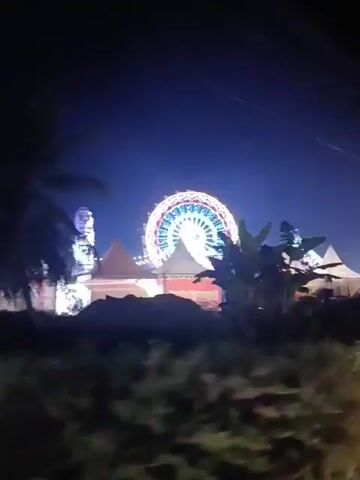 The video opens on a dark, grainy, night scene. In the background, a Ferris wheel is brightly illuminated with colorful lights, featuring a red, white, and blue circular pattern against a dark blue sky. To the left of the Ferris wheel, a tall, thin, white structure, possibly another amusement ride, is also lit up. In the middle ground, several white tents or canopies are visible, with some areas appearing to be stalls or booths. In the foreground, silhouetted dark shapes of foliage, likely trees and bushes, obscure the lower portion of the frame. The scene suggests an outdoor amusement park or festival at night, with the Ferris wheel being the dominant illuminated feature. The video is slightly shaky and appears to be filmed from a moving vehicle or by hand. Towards the end, two figures are partially visible in the bottom right corner, silhouetted against the lights. The overall impression is a lively, illuminated event taking place in the dark.