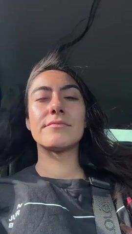 A young woman is depicted from the chest up, sitting in a vehicle in Imatra, Finland. Her long, dark hair is visibly blowing in the wind, suggesting either an open window or a convertible. She is wearing a black t-shirt with some partially visible white text, and a black seatbelt crosses her chest, featuring a white logo or text. Throughout the short clip, her eyes are mostly closed, and she maintains a serene and contented expression, occasionally opening them briefly. The lighting is bright and natural, indicating daytime, with sunlight illuminating her face. The car's interior appears dark, while the background outside the window is bright and blurred, hinting at motion. The woman seems to be enjoying the ride and the feeling of the wind.
