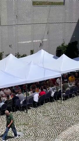The video shows an outdoor gathering with a long table set up for a meal. Several people are seated at the table, facing each other and engaged in conversation or eating. Above the table, a white tent provides shade. To the right of the table, a small musical ensemble is performing. The musicians consist of a man playing an upright bass, another playing an accordion, and a third person who appears to be playing a keyboard. They are set up on the cobblestone ground. In the background, there is a modern building with a metallic facade. A white van with lettering is parked near the entrance of the building. The overall scene suggests a festive event or a communal meal.
