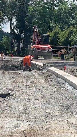The video shows a construction scene in La Fouillouse, France. The main subjects are a construction worker and an excavator. The worker, dressed in an orange high-visibility vest and trousers, is bending down and appears to be working on the ground. The excavator, a large red machine, is positioned behind the worker and is actively moving dirt or debris. The scene is set outdoors, with trees and foliage visible in the background, suggesting a daytime setting with clear weather. The ground is covered in gravel and dirt, typical of a construction site, with some freshly laid concrete curbs visible in the foreground. There are also traffic cones and other construction equipment present. A wooden fence runs along the back of the site. The overall impression is of active construction work. There is a logo on the excavator that reads "RECMAC".