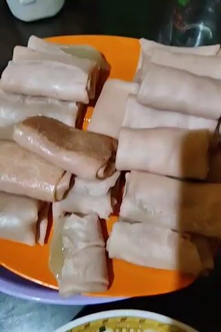Here's a description of the video:

The video shows a close-up of a plate of uncooked spring rolls. The plate is orange, and there are several rolls arranged on it. The spring rolls appear to be wrapped in a thin, pale skin. Some rolls look more translucent than others. The plate is sitting on a surface with a purple edge. Other items are partially visible in the background, including a clear glass and a white container.