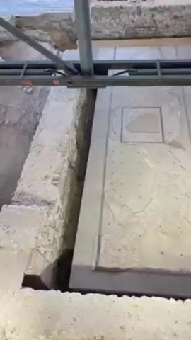 The video shows an archaeological dig, specifically a toilet from antiquity. The toilet consists of a stone slab with an opening in the center, and is surrounded by stone walls. There is a metal beam above it. The area around the toilet is excavated and the toilet itself is on a lower level. The slab has some cracks and missing pieces.  The person filming describes it.