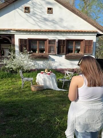 A young woman with long brown hair, wearing a white tank top and a white skirt, is engaged in taking photos or videos of an outdoor table setup in a garden in Rivića, Serbia. The scene is bathed in bright sunlight, casting distinct shadows.

In the background stands a charming white house with traditional wooden window frames and brown shutters. To the left of the house, a white-blossoming tree adds a touch of spring beauty. In the foreground, a small round table is draped with a flowing white tablecloth, set with several festive baked goods, some topped with red frosting, white eggs, and a vase filled with white flowers. Two grey chairs are positioned around the table, and a wicker basket rests on the grass nearby.

The woman is seen holding a smartphone, actively framing and capturing the scene. She reviews her shots on the screen, adjusts her position, and zooms in at various points, indicating a focused effort to get the perfect image or video. Her expression is concentrated as she interacts with her phone. The overall ambiance suggests a pleasant spring day, possibly centered around an Easter celebration given the presence of cakes and eggs. No discernible text is visible in the video.