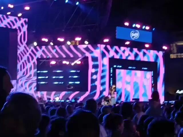 A wide shot captures a lively outdoor concert or event. A large stage dominates the background, adorned with vibrant LED screens displaying abstract, wavy patterns in shades of pink, purple, and blue. Multiple rows of stage lighting are visible, casting a colorful glow. A prominent blue banner with a white logo is displayed on one of the screens. In the foreground, a dense crowd of people is gathered, their backs mostly to the camera, suggesting they are facing the stage. The audience appears engaged, with some individuals looking towards the stage or interacting with each other. The overall atmosphere suggests a large-scale entertainment event.