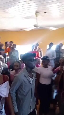 The video shows a crowded room, possibly a community hall, with people gathered for an event. A young woman wearing a tiara and a sash labeled "U.E.C." walks through the crowd, accompanied by a man in a cap and sunglasses. Other attendees are taking photos and videos with their phones. The room has yellow walls and a white ceiling with fans. Rows of blue chairs are visible in the background. The event seems to be celebratory or official, given the presence of the sash and tiara.