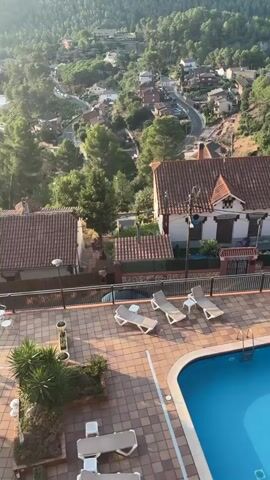 The video shows a high-angle view of a residential area nestled in a valley surrounded by lush green hills. In the foreground, a swimming pool area is visible with several empty lounge chairs on a tiled patio. Beyond the pool, rooftops of houses with terracotta tiles are scattered among the trees. A winding road can be seen snaking through the hillside, connecting the houses. The sun is casting a warm glow, indicating it might be early morning or late afternoon. The overall scene is serene and picturesque, highlighting a peaceful setting in nature.