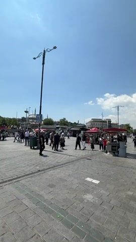 The video shows a bustling outdoor scene with many people walking and standing on a paved area. There are several food stalls and shops lining the right side of the frame, with tables and chairs set up outside some of them. Turkish flags are visible on top of some of the buildings. The sky is blue with some scattered clouds, and the sun is shining, casting shadows on the ground. In the distance, more buildings and structures can be seen. The overall atmosphere appears to be that of a busy public space, possibly a market or a waterfront area.