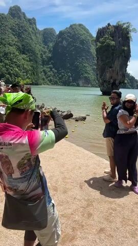 A man wearing a bright pink shirt with a bandana on his head and a bag slung across his body is taking a picture of a couple standing in front of a body of water. The couple is smiling and posing for the photo. In the background, there are large rock formations jutting out of the water, and the sky is blue with some clouds. Another man is visible in the water to the right of the couple. The scene appears to be a tourist destination.