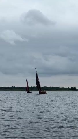 The video shows a body of water with several sailboats. The water is dark and choppy, and the sky is overcast with dark clouds. There are trees in the background, suggesting a landmass or shoreline. Two sailboats are clearly visible in the foreground and middle ground, with their sails up. One sailboat is closer to the camera and appears to be moving. Another sailboat is further away and also appears to be moving. The lighting suggests it is either early morning or late evening, or that the weather is poor. The overall mood is somber and calm.