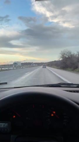 From the driver's seat of a car, the video shows a view of a highway with a white truck and a silver car ahead. The dashboard is visible, displaying warning lights. The video pans to the right, showing the passenger side mirror with raindrops on it, reflecting a view of the highway and the sunset. The sky above is partly cloudy. The video concludes with a close-up of the passenger side mirror.