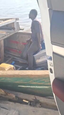 The video shows a man sitting on the motor of a small, wooden boat, operating it.  The boat is cluttered with pieces of wood, containers, and cloths. The video is taken from inside a vehicle, possibly a van or SUV. At one point, a Toyota Highlander with a Nigerian license plate (ABC31PQ) is visible on a wooden platform next to the boat. The environment appears to be a body of water, possibly a river or lake.