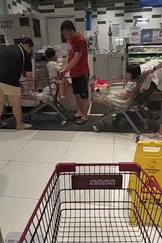 The video appears to be filmed in a supermarket. In the foreground, an empty shopping cart with a purple handle is visible. In the background, a family is shopping. A woman in a black shirt and beige shorts is placing items into a shopping cart. A man in a red and black shirt and black shorts is standing next to the cart, holding onto a child who is sitting in the cart. A second shopping cart with items inside is also visible. Further back, a woman in a green uniform is walking through the store, holding a white bag. Another section of the store shows a display of melons, with several shoppers browsing. The environment includes tiled flooring, refrigerated display cases, and shelving units stocked with products.