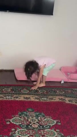 The video shows a young child, likely a toddler, standing on a rug in what appears to be an indoor setting. The child is wearing a light pink sleeveless shirt with "FASHION SPORTS" printed on it, along with cartoon bunny graphics. They are also wearing light mint green shorts. The child is barefoot.

In the foreground, a red rug with an intricate patterned design is visible. Behind the child, there are two pink cushions on the floor, and a pink object that may be a child's chair or toy is partially visible on the right. A white wall serves as the backdrop, and a dark television screen is mounted on the wall above. The lighting suggests it is daytime, possibly late afternoon due to the slightly dim lighting.

The child is actively engaged in an activity. They are holding a small, dark object, which they bring to their mouth and appear to be eating or about to eat. Their facial expression changes throughout the short clip, from looking curiously at the camera, to playfully holding the object near their eye, and finally to opening their mouth wide. The overall impression is one of playful curiosity and enjoyment. The child's hair is dark and somewhat tousled, suggesting movement or perhaps a windy day outside if this were an outdoor setting, but the interior setting makes it more likely due to the child's actions.