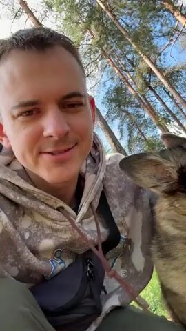 A man with short brown hair and wearing a patterned hooded sweatshirt is holding a medium-sized dog in his arms outdoors. The dog, which appears to be a mix of brown, tan, and gray fur, is close to the man's face. The man is talking to the camera and appears to be kissing or nuzzling the dog. The background shows a pine forest with trees reaching up towards a blue sky. The camera angle is low, looking up at the man and the dog.