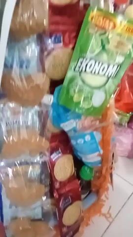 The video shows a tall, multi-tiered display of various packaged food items and what appear to be fresh produce, possibly for a celebratory occasion. The display is arranged in a cone-like structure. At the base, there are large green leaves, likely vegetables such as kangkung or bok choy, with additional items wrapped in clear plastic hanging down. Further up, there are multiple layers of packaged snacks, biscuits in clear plastic wrappers, and some red-colored packets. A prominent green pouch of dish soap is also visible, attached to the side of the display. The top of the structure appears to be adorned with stacked clear plastic containers filled with more biscuits or snacks. In the background, other stalls or displays are visible, along with some people whose faces are not clearly discernible. The overall impression is of a market stall or a display set up for a special event, possibly a bazaar or a religious festival like Eid al-Fitr, given the presence of the crescent moon and lantern imagery on one of the packages in the lower left. The lighting suggests an indoor environment, possibly a covered market or shop.