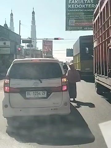 The video shows a street scene with several vehicles and a pedestrian. In the foreground, a white minivan is stopped, facing away from the camera. Its license plate reads "BL 1254 ZF". To the right of the minivan, a person wearing a headscarf and a long, dark skirt is walking away from the camera. Behind the minivan, a large truck is visible. Further in the background, there are buildings, including a tall white structure that appears to be a minaret, and a green billboard with text in Indonesian, including "FAKULTAS KEDOKTERAN" (Faculty of Medicine). Traffic lights are visible, and one appears to be red, indicating that traffic is stopped. The sky is clear and the sun is shining, casting shadows on the road. The overall impression is a typical urban street scene during the daytime.