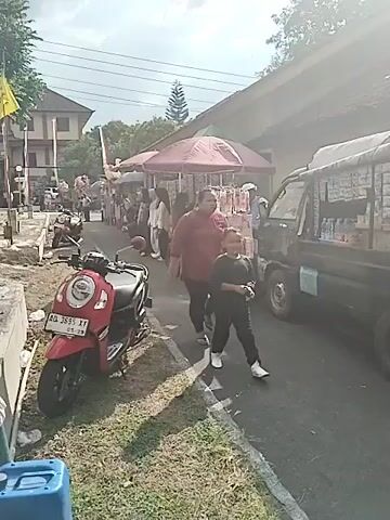 Here is a description of the video:

The video shows an outdoor event taking place in a public space. The setting appears to be a local gathering or celebration. There are rows of vendors selling various items along a street, indicated by parked vehicles. There are several people walking through the area, including a woman and a child walking in the foreground. Some are looking at the vendors, while others are walking through. 

There is also a designated seating area with green plastic chairs under a tent or awning. A crowd of people are seated in the chairs. The area seems set up for a performance or presentation, as there is a stage and a screen visible in the background. People are milling around in the seating area, suggesting that the event is about to begin or is ongoing. The weather appears sunny, and the atmosphere suggests a community event or festival.