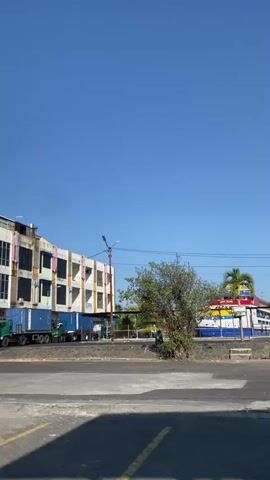 A clear, blue sky dominates the upper portion of the video. In the foreground, a grey asphalt road with yellow painted lines is visible, and a portion of a black and teal motorcycle is parked on the right. The road leads to a paved area with interlocking grey bricks. Several red and white striped flags on poles are prominent, with two larger flags on the right side of the frame. A yellow barrier with red and white striped tape is positioned in front of the flags. A building with a dark-tiled roof and a blue and orange facade is partially visible on the left. Further back, a multi-story building with an unfinished facade, featuring concrete and some yellow framing, is present. Several shipping containers are stacked near this building, and a green truck is parked to the left of the containers. A small tree with sparse green leaves stands in the middle ground, positioned between the buildings and the water. In the background, a large ferry, painted blue, red, yellow, and white, is docked. Palm trees are visible behind the ferry. The overall scene appears to be a port or ferry terminal on a sunny day.