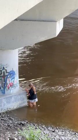 A woman is standing in muddy river water under a concrete bridge. The bridge is covered in graffiti. The woman is wearing a black t-shirt and gray shorts, and her hair is in a braid. The river water is brown and appears to be flowing. Sunlight is creating a reflection on the water.