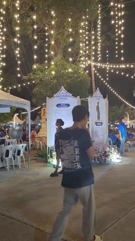 The video shows an outdoor night scene decorated with numerous strings of small, warm white lights hanging from trees. In the foreground, a paved walkway is visible. To the left, a white, peaked tent is set up with a table and white plastic chairs in front of it. Several people are positioned behind the table, and some appear to be working. A large industrial fan is also present near the tent. Further into the background, more tents and structures are visible, along with more people. In the center of the frame, two large informational displays on stands are set up, adorned with floral arrangements at their bases. The display on the right features text in a foreign language and images. Several people are walking through the area. One person, wearing a dark t-shirt with a graphic design and light-colored pants, is walking away from the camera in the foreground. Another person, wearing a patterned dress, is walking towards the informational displays. The overall atmosphere suggests a lively outdoor event or market at night.