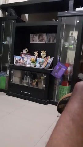 The video shows a stationary view of a dark-colored media console with glass-fronted cabinets. The console is filled with various items, including framed photographs, decorative objects, and possibly trophies. A purple binder is leaning against the right side of the central shelf. To the left of the console, a green plastic bag is visible on the floor. The floor appears to be tiled. A portion of a person's leg and foot, wearing what looks like a brown shoe, is visible in the lower right corner of the frame. Curtains are visible on the far left of the scene.