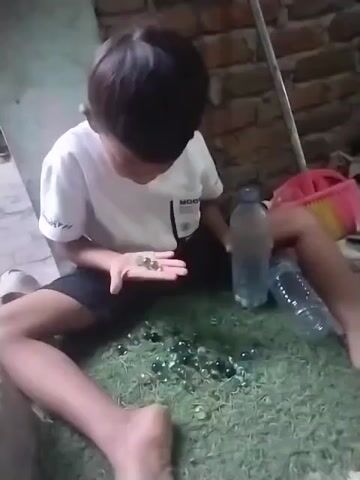 The video shows a young boy with dark hair and a white t-shirt sitting on a green surface, possibly artificial grass. He is holding a few marbles in his left hand and looking down at them. Scattered in front of him on the green surface are numerous marbles of various colors, predominantly green and clear. To his right, there are two plastic water bottles, one upright and the other lying on its side. The background consists of a brick wall and some other indistinct objects. The boy appears to be playing with the marbles.