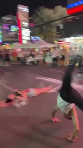 The video shows a male performer putting on a show for a crowd on a street at night. The performer, wearing a white tank top and black pants, is dancing and throwing a set of sharp knives in the air, and then stabbing the ground as he dances around a woman lying on the ground on a striped mat. The woman is wearing a light tank top and shorts and her head is covered with a small clear plastic box. The crowd watches from the edge of the street in front of a colorful array of shops and market tents. Bright lights illuminate the scene.