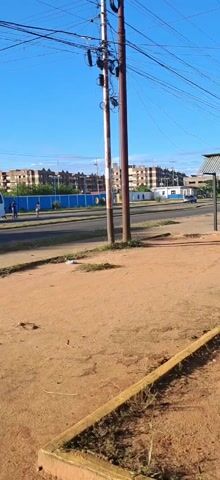 The video depicts an urban scene during daylight.  In the foreground is a dirt area, bordered by a short concrete curb. Two utility poles with multiple wires are centered.  Beyond is a road with vehicular traffic, including a white box truck and motorcycles. Across the road is a blue fence and low buildings, with larger apartment-style buildings further in the distance.  A bus shelter stands to the right. A few pedestrians are visible near the blue fence.  The sky is blue and mostly clear.