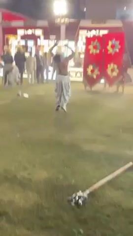 At night, on a grassy lawn, a shirtless man wearing white pants performs gymnastic moves in front of a small crowd.  He completes two backflips, and there are decorative spears on the ground near him. The crowd of people, many in turbans, stand in the background watching.  Behind the crowd is a brightly lit event space and buildings. The lawn is illuminated with spotlights.