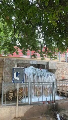 The video shows a man-made waterfall cascading down a stone and tile structure. Lush green leaves from a tree overhang the top of the frame. In front of the waterfall, there is a metal fence with decorative elements. To the left of the waterfall, a blue sign with white writing and an 'F' is attached to the structure. The overall scene appears to be outdoors, possibly in a park or a public space. The water flow is continuous and appears to be strong.