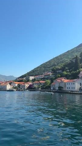 A video shows a coastal town with buildings on a hill overlooking a body of water. The buildings are of various sizes and colors, with red tile roofs being prominent. Trees and lush green vegetation cover the hillside. The water in the foreground is blue and appears to have some floating debris. The sky is clear and blue. The camera appears to be moving slightly, suggesting it is on a boat or in a moving vehicle.