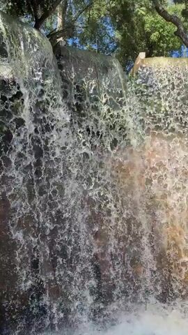 A vertical close-up shot displays water cascading down a tiered artificial waterfall. The water is clear and bubbly as it hits the bottom pool. Some greenery is visible at the top of the frame, suggesting nearby trees. The light is bright and causes reflections on the water surface.