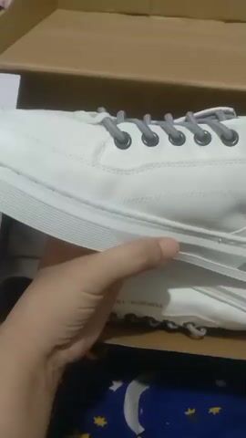 The video shows a person holding a white sneaker with gray laces. The sneaker is being taken out of a cardboard box. The camera pans across the sneaker, showing its side, laces, and sole. The person's hand is visible in the foreground. The background shows part of the box and a blue and yellow patterned fabric. The lighting is dim.