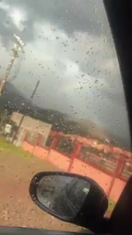 The video, shot from inside a car, shows a view of a town or settlement nestled in a mountainous landscape under a cloudy sky. Raindrops are visible on the car window, suggesting recent or ongoing precipitation. A partial rainbow arcs across the sky in the distance, positioned above the settlement. In the foreground, the car's side mirror, reflecting the rainy environment, is visible. A power line with poles is also seen running alongside the road. The settlement appears to have buildings with red accents.