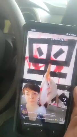 The video shows a person holding a smartphone. The smartphone displays a social media video. In the social media video, there are two frames. The top frame shows an abstract painting with black and red markings. A person in white clothing, with dramatic makeup and black hair, is positioned in front of the painting, their arms outstretched. The bottom frame of the social media video shows a selfie of a person with short brown hair and a surprised expression, looking directly at the camera. The caption on the social media video reads "Her portrait is much better than I usually do". The bottom of the smartphone screen shows interface elements for commenting and liking. The background of the real-world footage shows the interior of a car.