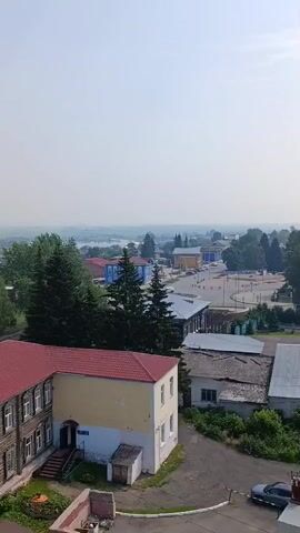 The video captures an aerial view of a town. In the foreground, a two-story building with a red roof and wooden siding is visible on the left. Next to it, a yellow and white building with a flat roof has a blue sign above its entrance. Further back, a wider view shows a town square with a few buildings and a street. Trees are scattered throughout the scene, and a body of water can be seen in the distance. The sky is hazy, suggesting smog or dust. A dark-colored car is parked on the right side of the road in the foreground.