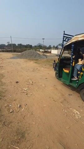 The video shows a green auto-rickshaw stationary on a dirt lot. The vehicle has a rack on top, and advertisements on its side panels. A person wearing jeans is seated inside. Behind the auto-rickshaw is a pile of gravel. The ground is a mix of dirt and debris. The sky is clear and blue. In the distance, there are trees and power lines. The video shows the rickshaw from the side, then the view pivots to show the back of the vehicle. The engine is running.