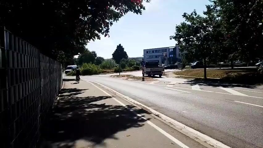 The video captures a sunny day on a street with a bicycle lane running along a fenced area. A person on a bicycle is visible in the distance in the bicycle lane. A white van drives on the road, approaching a roundabout. In the background, a modern building complex is visible with cars parked in front. Trees line the street, casting shadows on the bicycle lane.