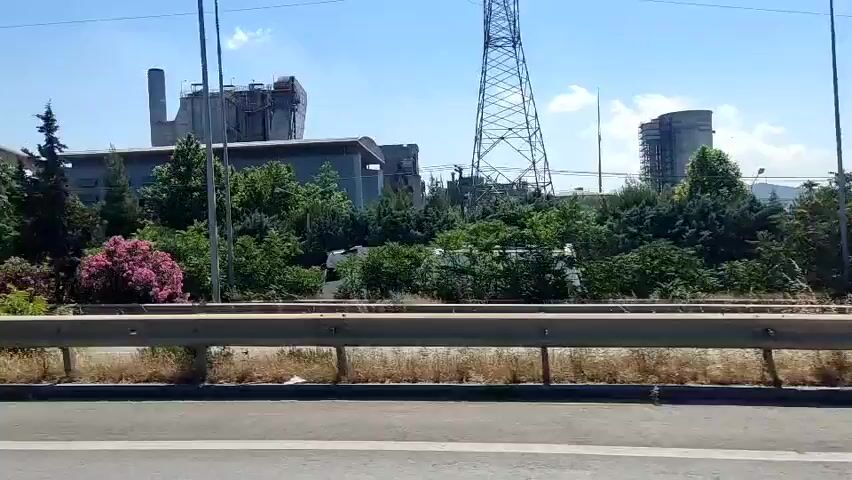 The video displays an outdoor scene on a clear, sunny day, captured from a moving vehicle traveling parallel to a road. In the foreground, a metal guardrail separates the camera's perspective from a multi-lane asphalt road. Beyond the road, a strip of green foliage, including trees and bushes with some pink flowers, lines the edge of an industrial complex. The complex features large gray and blue industrial buildings, including tall silos, structures resembling a cement factory with a visible conveyor belt system, and multiple chimneys. A tall metal lattice tower is visible on the right side of the frame. At 0:02, a dark gray Audi station wagon drives from right to left across the road in the foreground. The camera pans steadily from right to left throughout the video, revealing more of the industrial landscape.