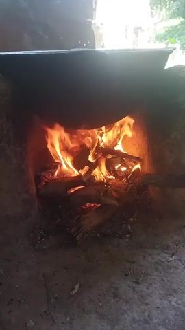 A fire burns in a clay oven. A large pot sits on top of the oven, obscuring the view of what is inside. A hand enters the frame and uses a stick to adjust the burning logs in the oven. The scene is dimly lit, with the fire casting an orange glow on the surrounding dirt and clay. Lush green foliage is visible in the background, suggesting an outdoor setting.
