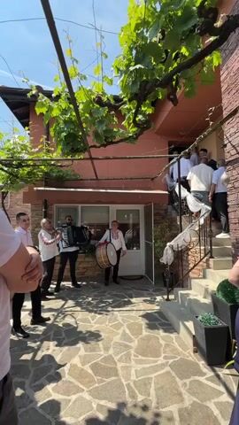 A medium shot captures a band of six men playing instruments in a courtyard. The men wear a mix of white shirts and patterned shirts. One man plays an accordion, another a drum, and others play wind instruments. A grape vine with green leaves covers a trellis above them. In the background, a two-story terracotta-colored house has an open door, revealing a group of men standing on a staircase. The house has white trim around its windows and a white railing on the stairs. In the foreground, the lower bodies of two people on the left and right are visible, observing the band. The ground is paved with irregular stone slabs. The scene is brightly lit by the sun, casting shadows on the ground.