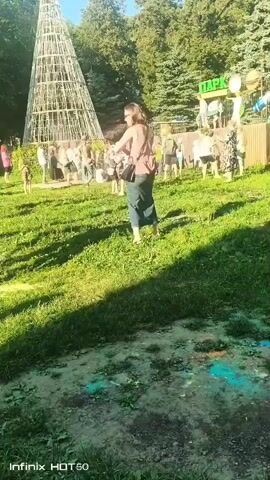 A group of people are gathered in a park, some are throwing colored powder at each other. In the foreground, a child in a red shirt and a woman in a brown shirt and green skirt are interacting. Behind them, there are trees, a large conical structure made of wood, and a sign that says "ПАРК". The ground in the foreground is stained with colored powder. The video appears to be taken on a sunny day.