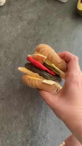 A hand is seen playing with a fabric toy hamburger. The hamburger is made of different fabric pieces representing the bun, patty, cheese, tomato, and pickles. The hand picks up the top bun and then separates the layers of the hamburger. The background is a dark, textured surface, possibly a countertop.