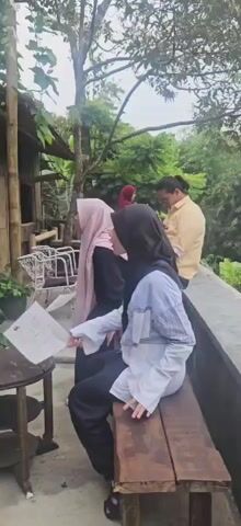 A group of people are gathered outdoors. In the foreground, a woman wearing a black hijab and a light blue and white patterned shirt is seated on a wooden bench, holding a piece of paper. To her left, another woman in a pink hijab is also seated and holding paper. In the background, a person in a yellow shirt stands, looking down at something. To the far left, there are white chairs and a thatched-roof structure. The environment appears to be a natural setting with trees and foliage.
