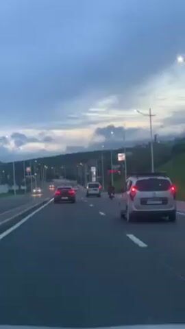 A medium shot captures traffic on a multi-lane road during the twilight hours, under a sky streaked with clouds. Several cars, including a silver Dacia Lodgy with roof racks and visible license plate "55054", and a red hatchback, move along the road. A motorcyclist, wearing red, is seen splitting lanes. Streetlights illuminate the roadside. In the distance, signs are visible, hinting at commercial establishments. The scene is generally calm and orderly, depicting everyday urban traffic at dusk.