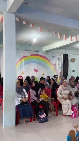 The video shows a group of people, mostly women and children, seated on red chairs in an indoor room. A young woman in a white vest and black shirt is standing in the foreground, looking towards the camera with her mouth open. The room is decorated with a rainbow banner and small red and white flags. Some of the people in the audience are smiling and looking at the young woman. The scene appears to be a performance or event for children.